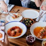 Close-up of a restaurant table with a variety of dishes as a group of friends takes pictures of the delicious food with their phones.