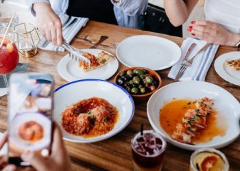 Close-up of a restaurant table with a variety of dishes as a group of friends takes pictures of the delicious food with their phones.