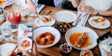 Close-up of a restaurant table with a variety of dishes as a group of friends takes pictures of the delicious food with their phones.