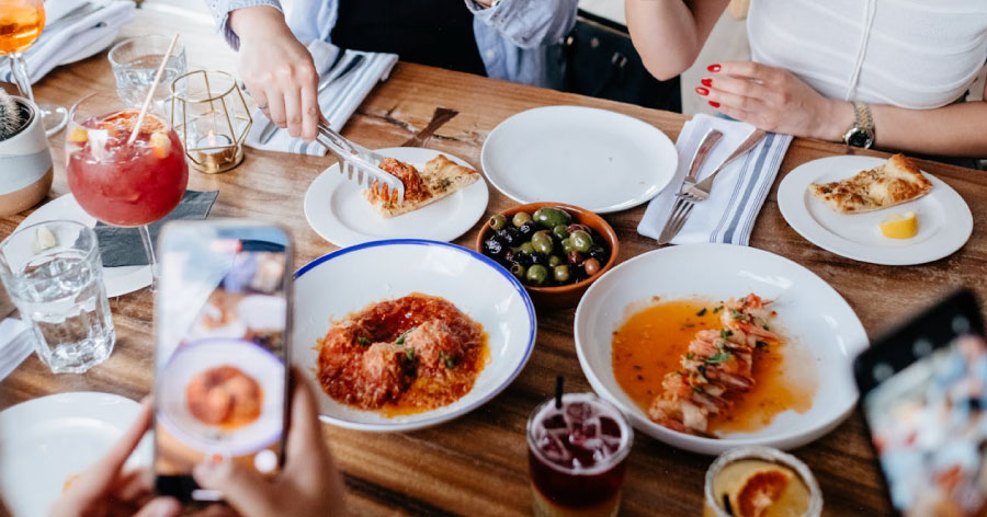 Close-up of a restaurant table with a variety of dishes as a group of friends takes pictures of the delicious food with their phones.