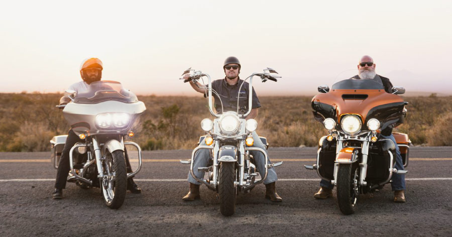 Three men are sitting side by side on their motorcycles with their headlights on, posing for a photo on the side of the road.