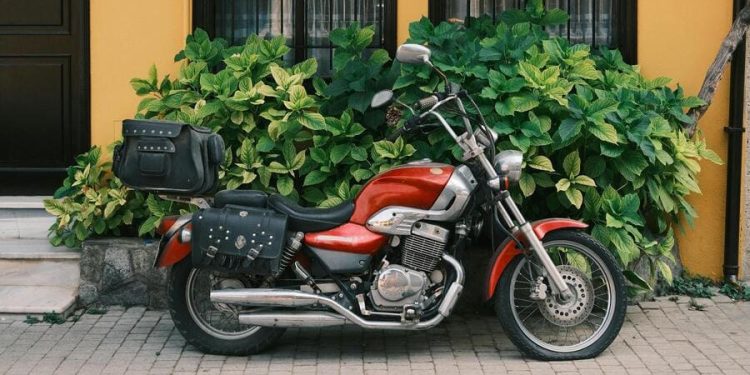 A red cruiser motorcycle parked in front of a yellow house with black trim and lush greenery in a quiet residential neighborhood.