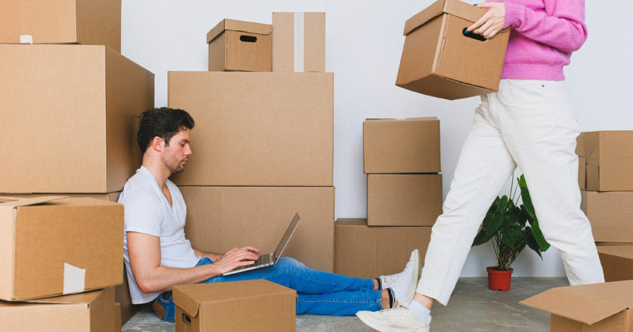 A woman is carrying a box past a man who is sitting on the floor, leaning up against a stack of boxes while researching small business storage solutions on his laptop.
