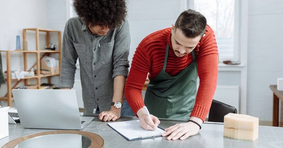 Two small business owners are reviewing documents and product plans at a worktable beside a laptop with small business storage shelves in the background.