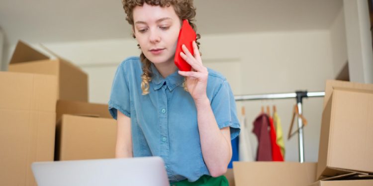 A woman is leaning over her laptop while talking on her cellphone, surrounded by cardboard boxes because she hasn’t found the right small business storage solution.