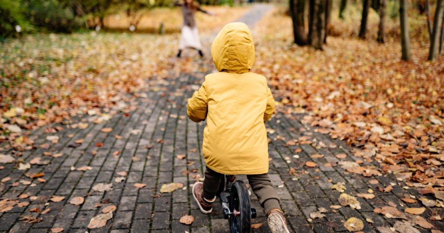 A small child in a yellow coat is riding a bike along a park path in the fall.