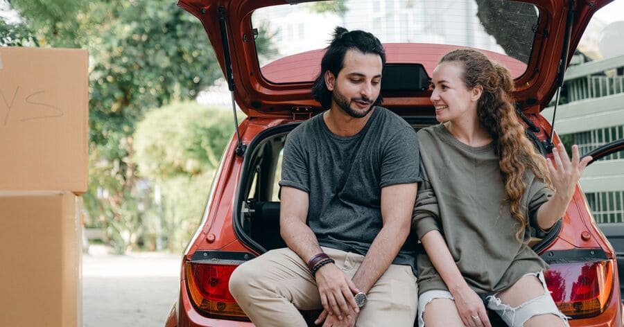 A couple is resting against the rear bumper of their car after moving to Charlotte, North Carolina.