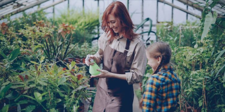 A woman with red hair is showing a young girl how to water plants with a spray bottle and other greenhouse gardening tips.