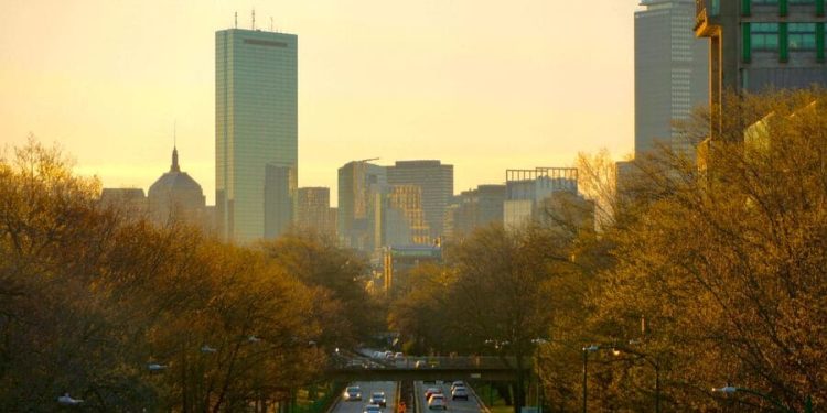 The Boston skyline rises above a tree-lined boulevard at sunset as cars commute in and out of the city center.
