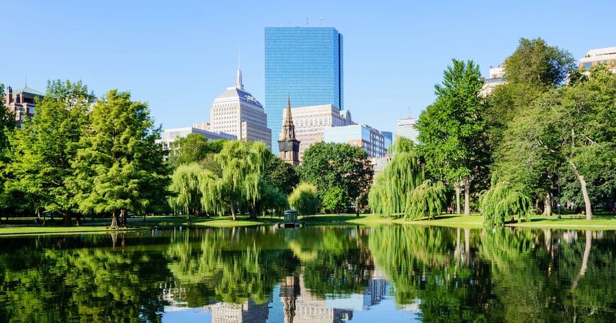 View of the Boston skyline rising above Boston Common, with lush green trees reflected in a calm pond on a sunny day.
