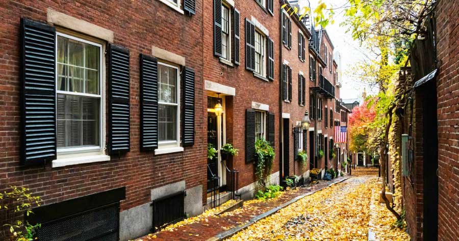 View down a quaint side street in Beacon Hill, featuring brick townhomes with black shutters and sidewalks littered with autumn leaves.