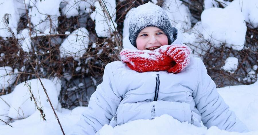 A child is bundled in a white winter coat, knit hat, and red scarf, happily playing in deep snow on a winter day.