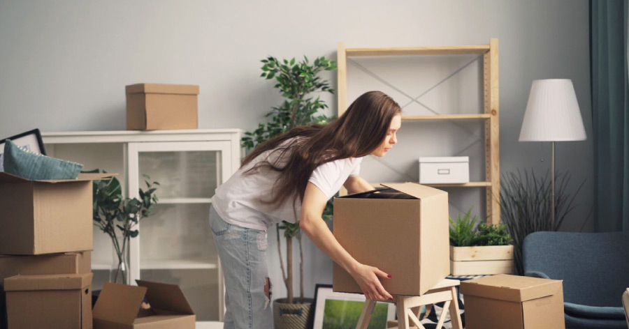 A woman is bending over to pick up a moving box from a step stool while in the process of moving to Boston.