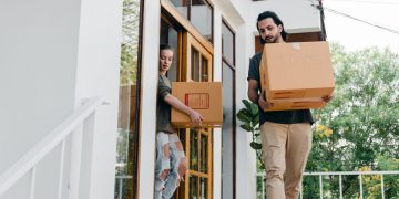 Two people carry moving boxes out of their home after labeling them and clearing a path; simple tips for moving in the summer.