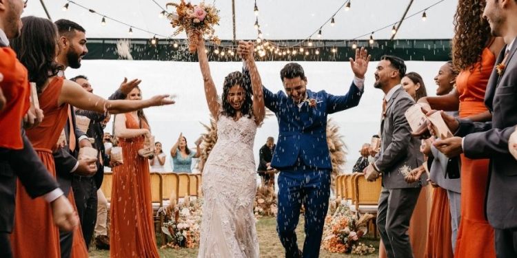 A newly married couple is walking hand in hand down the aisle after saying “I do” as their friends and family throw rice in the air and cheer them on.