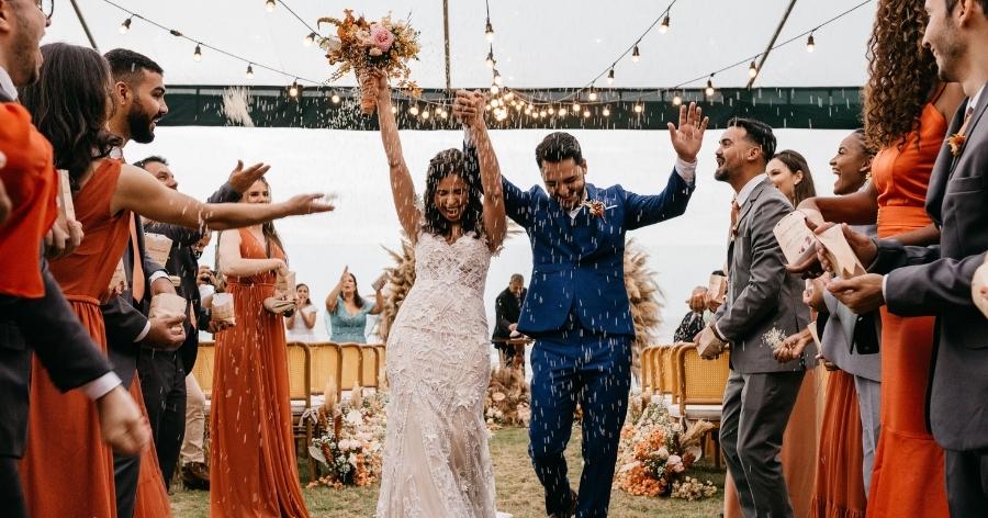 A newly married couple is walking hand in hand down the aisle after saying “I do” as their friends and family throw rice in the air and cheer them on.