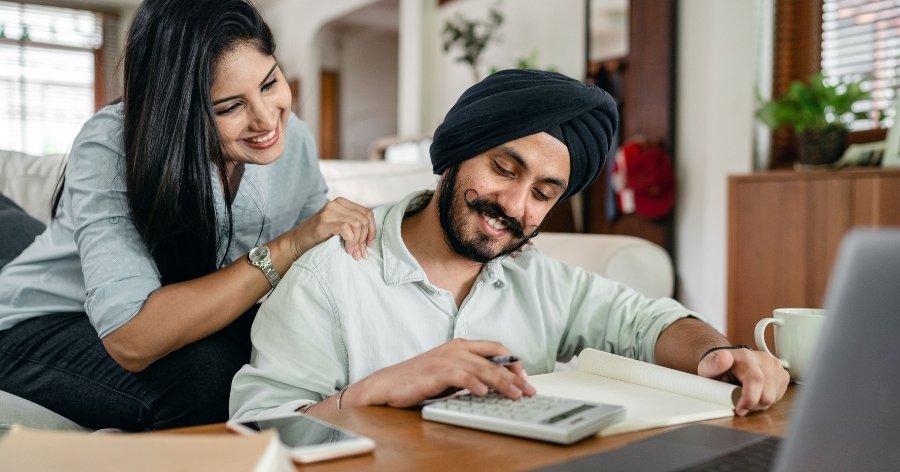 A happy couple is sitting in their living room, calculating their budget while DIY wedding planning.