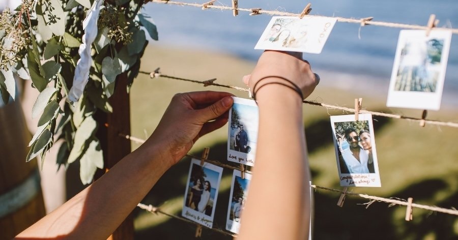 A pair of hands is using small wooden clothespins to hang instant photos of the happy couple and their friends on lengths of twine, a clever idea for DIY wedding decorations.