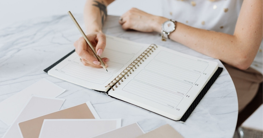A woman is writing her DIY wedding planning checklist in a planner at her dining table.